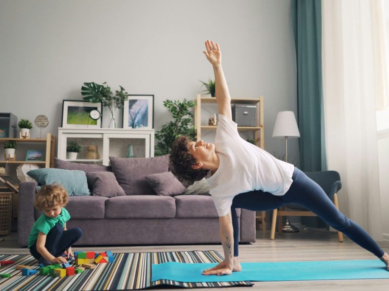 Frau macht Yoga auf einer Matte, während ein Kind mit Bauklötzen spielt.