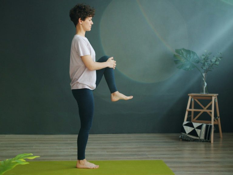 Eine Person steht auf einem Yoga-Matte und hebt ein Knie zur Brust.