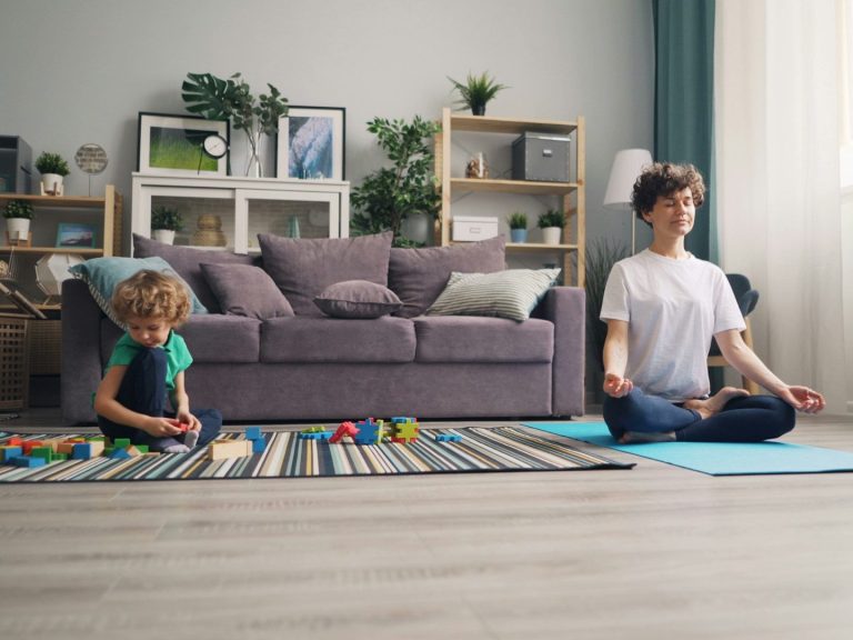 Frau meditiert im Wohnzimmer, während ein Kind mit Bauklötzen spielt.